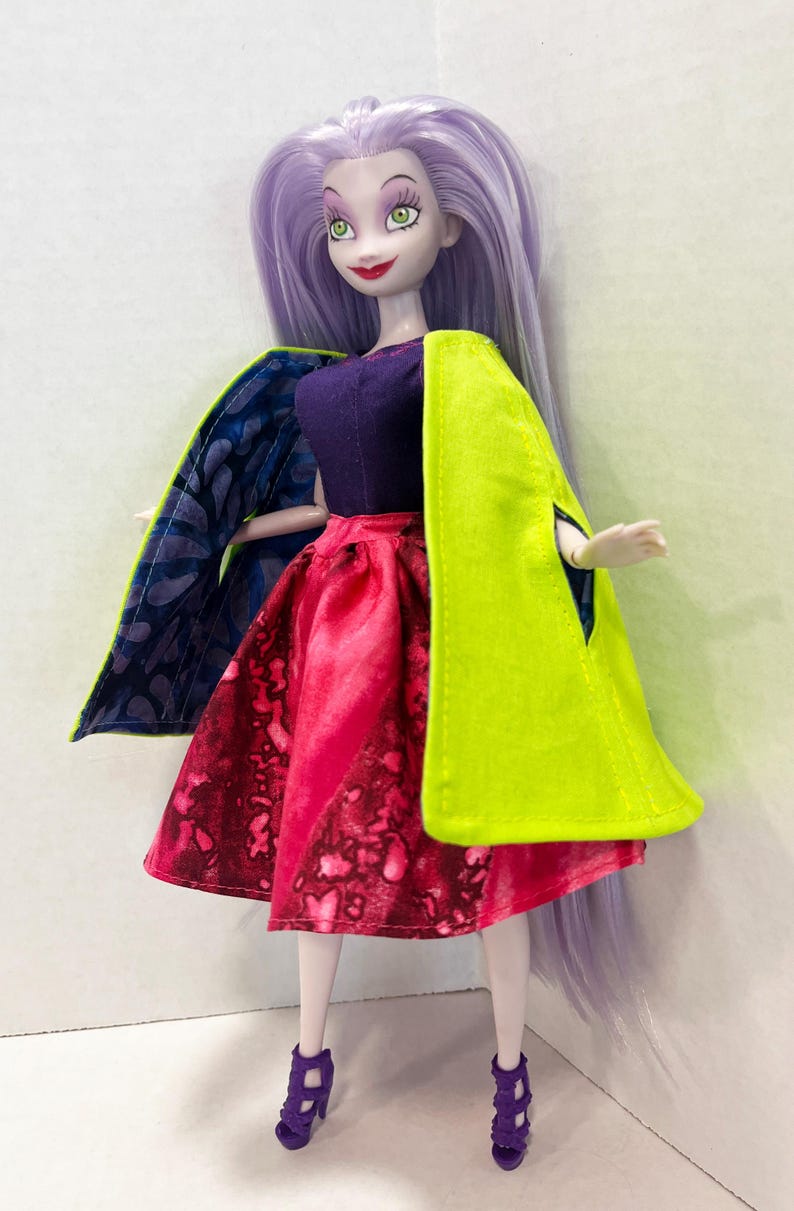 Pu&ograve; includere: Una bambola con lunghi capelli color lavanda, occhi verdi e labbra rosse. La bambola indossa un top viola scuro, una gonna con motivo rosso e rosa, un mantello verde lime brillante con fodera a motivi blu e viola e tacchi viola.