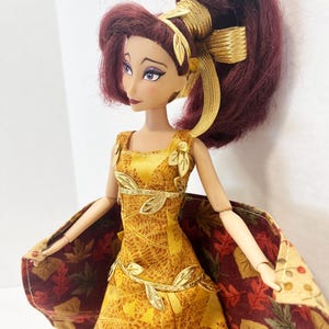 May include: A doll with dark red hair styled in a high ponytail with a gold ribbon. The doll wears a yellow dress featuring a gold leaf pattern, gold leaf trim at the waist and shoulders, and a cape with a multi-colored autumn leaf print pattern.