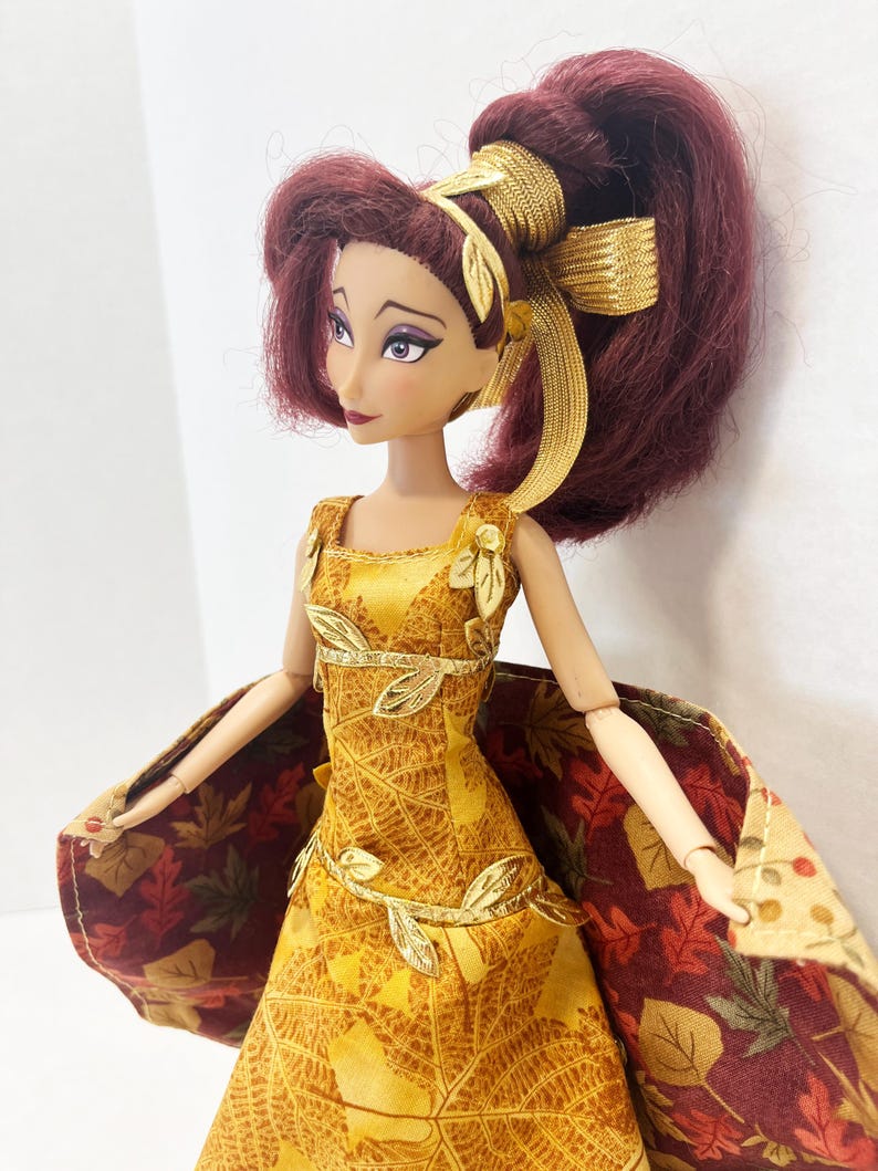May include: A doll with dark red hair styled in a high ponytail with a gold ribbon. The doll wears a yellow dress featuring a gold leaf pattern, gold leaf trim at the waist and shoulders, and a cape with a multi-colored autumn leaf print pattern.