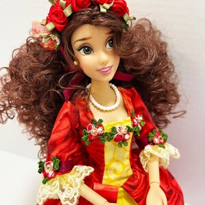 May include: A doll with brown curly hair and green eyes, wearing a red dress with a rose pattern and yellow accents. The doll has a red rose crown and a pearl necklace. The dress has lace trim and floral embellishments.