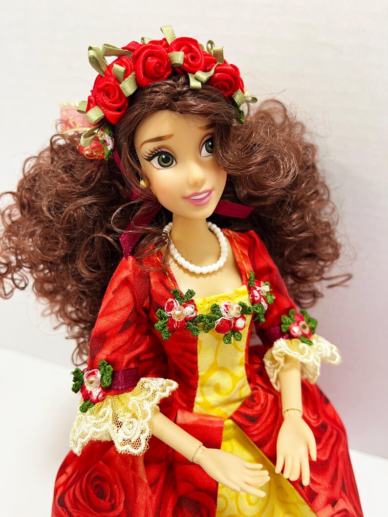 May include: A doll with brown curly hair and green eyes, wearing a red dress with a rose pattern and yellow accents. The doll has a red rose crown and a pearl necklace. The dress has lace trim and floral embellishments.