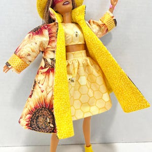Puede incluir: Una mu&ntilde;eca vestida con un conjunto amarillo, con un abrigo con estampado de girasoles y un sombrero a juego. El conjunto incluye un top corto y una falda con estampado de panal y detalles de abejas. La mu&ntilde;eca lleva botas amarillas con suelas rosas.