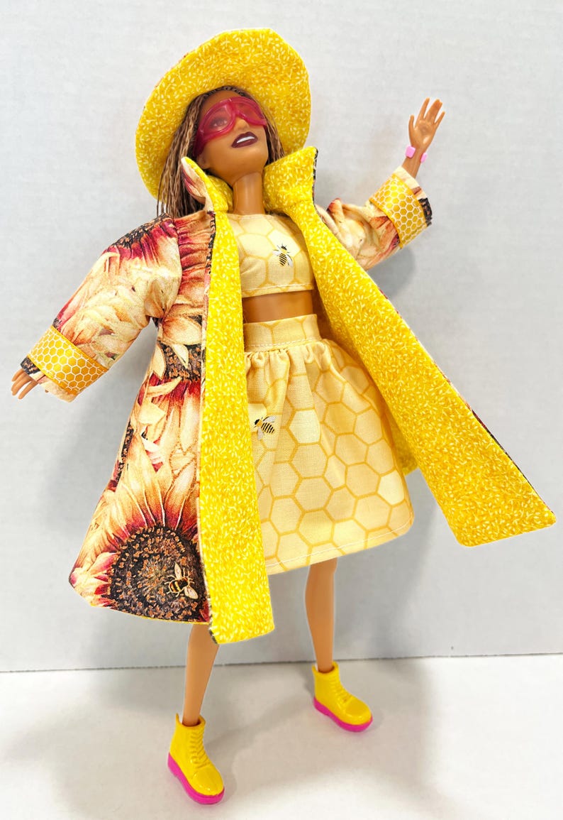 Puede incluir: Una mu&ntilde;eca vestida con un conjunto amarillo, con un abrigo con estampado de girasoles y un sombrero a juego. El conjunto incluye un top corto y una falda con estampado de panal y detalles de abejas. La mu&ntilde;eca lleva botas amarillas con suelas rosas.