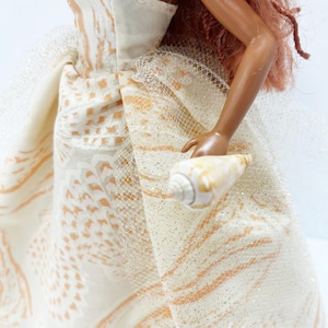 Puede incluir: Una mu&ntilde;eca con un vestido crema sin tirantes con un patr&oacute;n de formas naranjas y blancas. El vestido tiene una superposici&oacute;n de tul con purpurina dorada. La mu&ntilde;eca sostiene una concha marina.