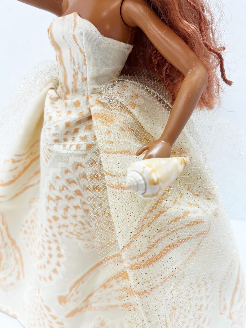 Puede incluir: Una mu&ntilde;eca con un vestido crema sin tirantes con un patr&oacute;n de formas naranjas y blancas. El vestido tiene una superposici&oacute;n de tul con purpurina dorada. La mu&ntilde;eca sostiene una concha marina.