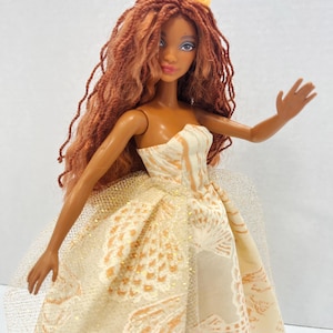 Puede incluir: Una mu&ntilde;eca de moda con cabello largo color casta&ntilde;o rojizo y una corona amarilla. La mu&ntilde;eca lleva un vestido crema sin tirantes con un estampado dorado y blanco. La mu&ntilde;eca tiene la piel oscura, ojos azules y una ligera sonrisa.