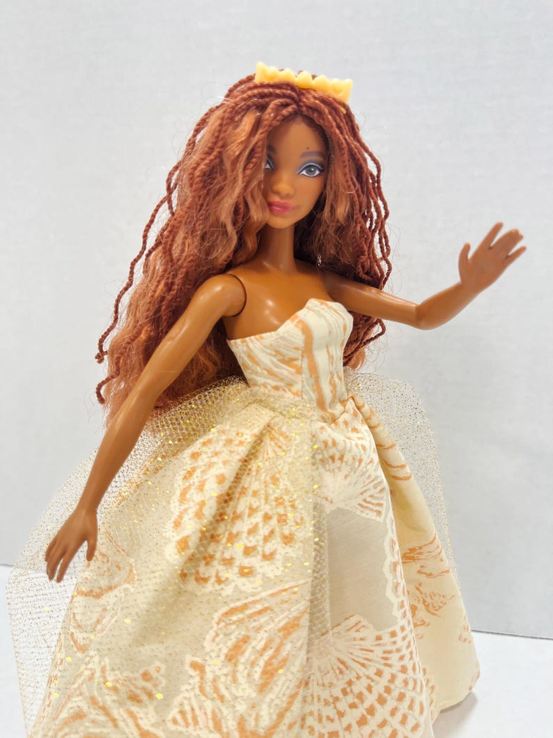 Puede incluir: Una mu&ntilde;eca de moda con cabello largo color casta&ntilde;o rojizo y una corona amarilla. La mu&ntilde;eca lleva un vestido crema sin tirantes con un estampado dorado y blanco. La mu&ntilde;eca tiene la piel oscura, ojos azules y una ligera sonrisa.