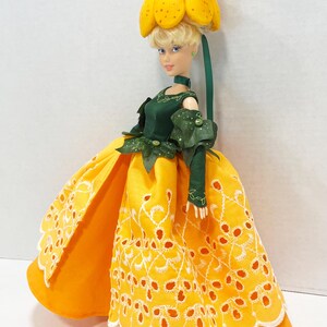 May include: A doll with blonde hair wearing a yellow flower-shaped hat and a dark green bodice with matching sleeves. The doll wears a long, bright yellow skirt featuring a white eyelet lace overlay with a scalloped hem and floral embroidery.