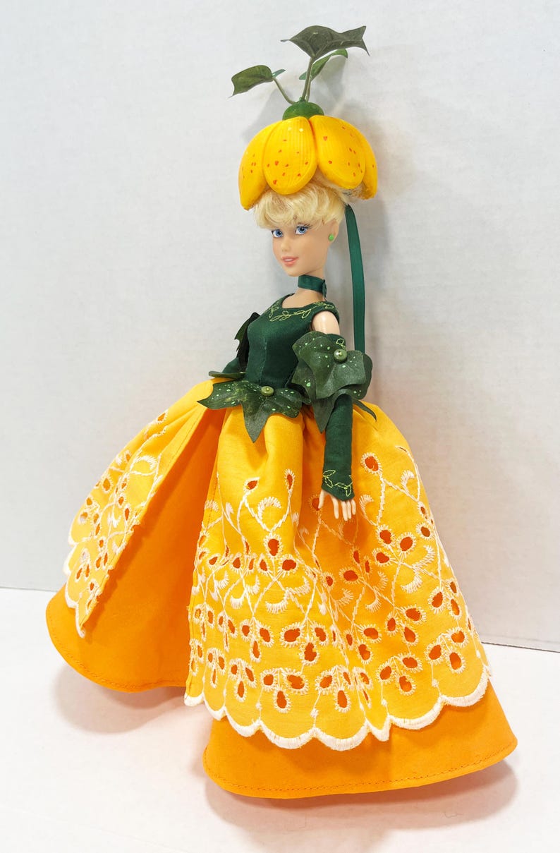 May include: A doll with blonde hair wearing a yellow flower-shaped hat and a dark green bodice with matching sleeves. The doll wears a long, bright yellow skirt featuring a white eyelet lace overlay with a scalloped hem and floral embroidery.