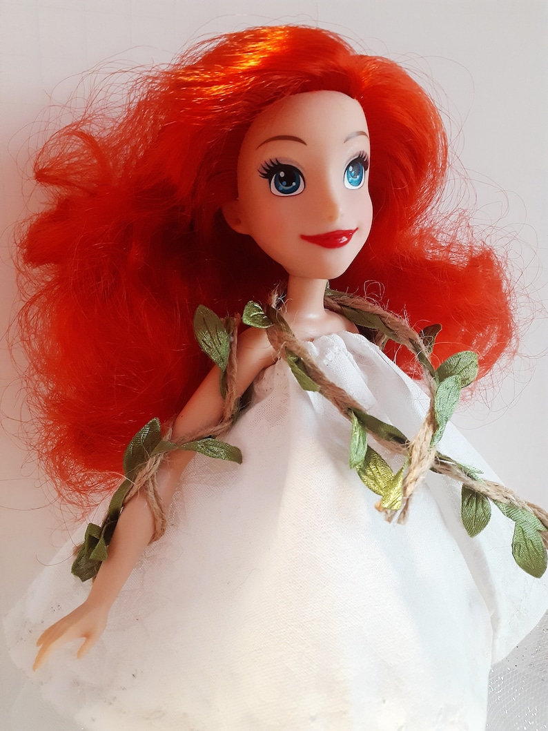 Puede incluir: Una mu&ntilde;eca de pelo rojo con un vestido blanco con hojas verdes y cuerda envuelta alrededor.