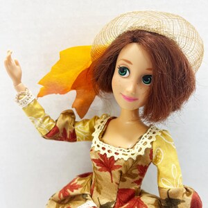 May include: A doll with short auburn hair, green eyes, and freckles, wearing a dress with a brown and red maple leaf pattern, yellow sleeves, and white lace trim. The doll has a tan mesh hat and a large orange fabric leaf attached to the side.