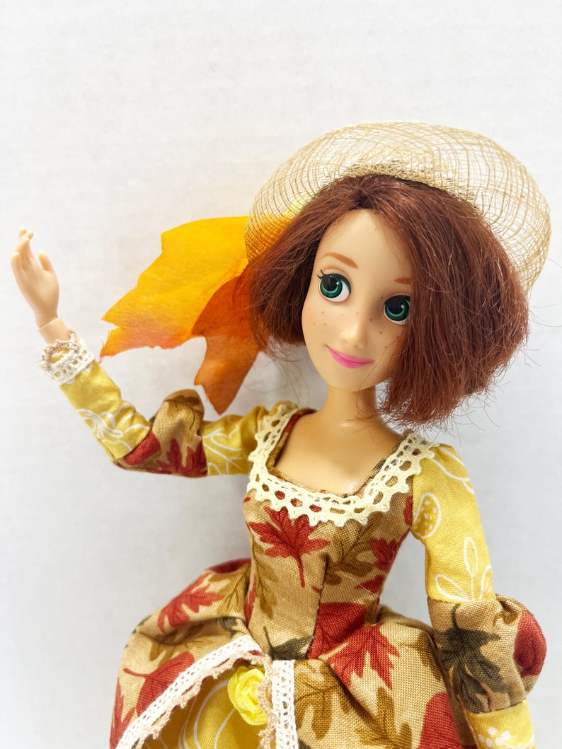 May include: A doll with short auburn hair, green eyes, and freckles, wearing a dress with a brown and red maple leaf pattern, yellow sleeves, and white lace trim. The doll has a tan mesh hat and a large orange fabric leaf attached to the side.