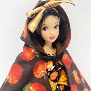 Op de afbeelding: Een pop met zwart haar draagt een bruine cape met capuchon met een rode appelprint en een beige geruite strik. De pop heeft bruine ogen, rode lippen en draagt een zwart kledingstuk met een patroon van appelschijfjes en een geel accent.