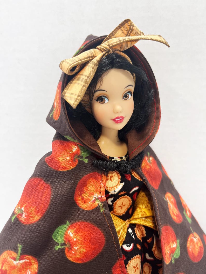 Op de afbeelding: Een pop met zwart haar draagt een bruine cape met capuchon met een rode appelprint en een beige geruite strik. De pop heeft bruine ogen, rode lippen en draagt een zwart kledingstuk met een patroon van appelschijfjes en een geel accent.