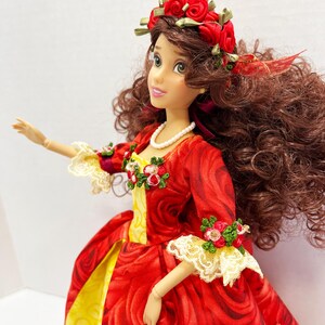 May include: A doll wearing a red rose-patterned dress with yellow accents and a white lace trim. The doll has a red rose crown and a pearl necklace. The doll has brown curly hair and is posed with one arm extended.
