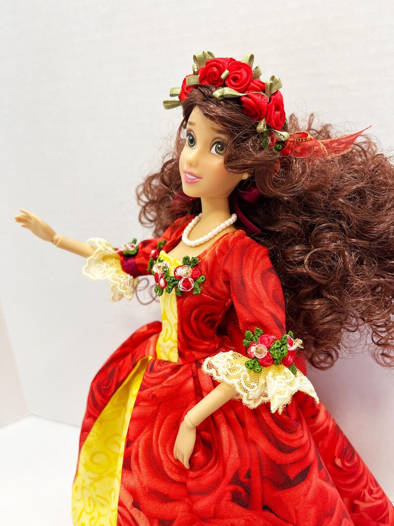 May include: A doll wearing a red rose-patterned dress with yellow accents and a white lace trim. The doll has a red rose crown and a pearl necklace. The doll has brown curly hair and is posed with one arm extended.