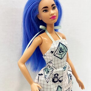 May include: A doll with long, vibrant blue hair and dark eyebrows, wearing a white halter-neck romper featuring a black grid pattern and various gaming dice illustrations, including a prominent ampersand symbol and the number eighteen.