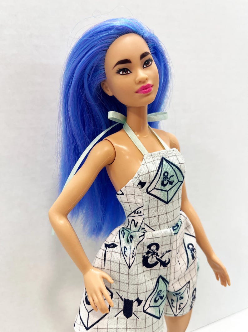 May include: A doll with long, vibrant blue hair and dark eyebrows, wearing a white halter-neck romper featuring a black grid pattern and various gaming dice illustrations, including a prominent ampersand symbol and the number eighteen.