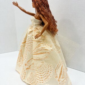 Puede incluir: Mu&ntilde;eca con largo cabello casta&ntilde;o rojizo y un vestido sin tirantes color crema. El vestido presenta un estampado de conchas en tonos dorados. Una tela transparente y brillante se extiende desde la espalda del vestido.