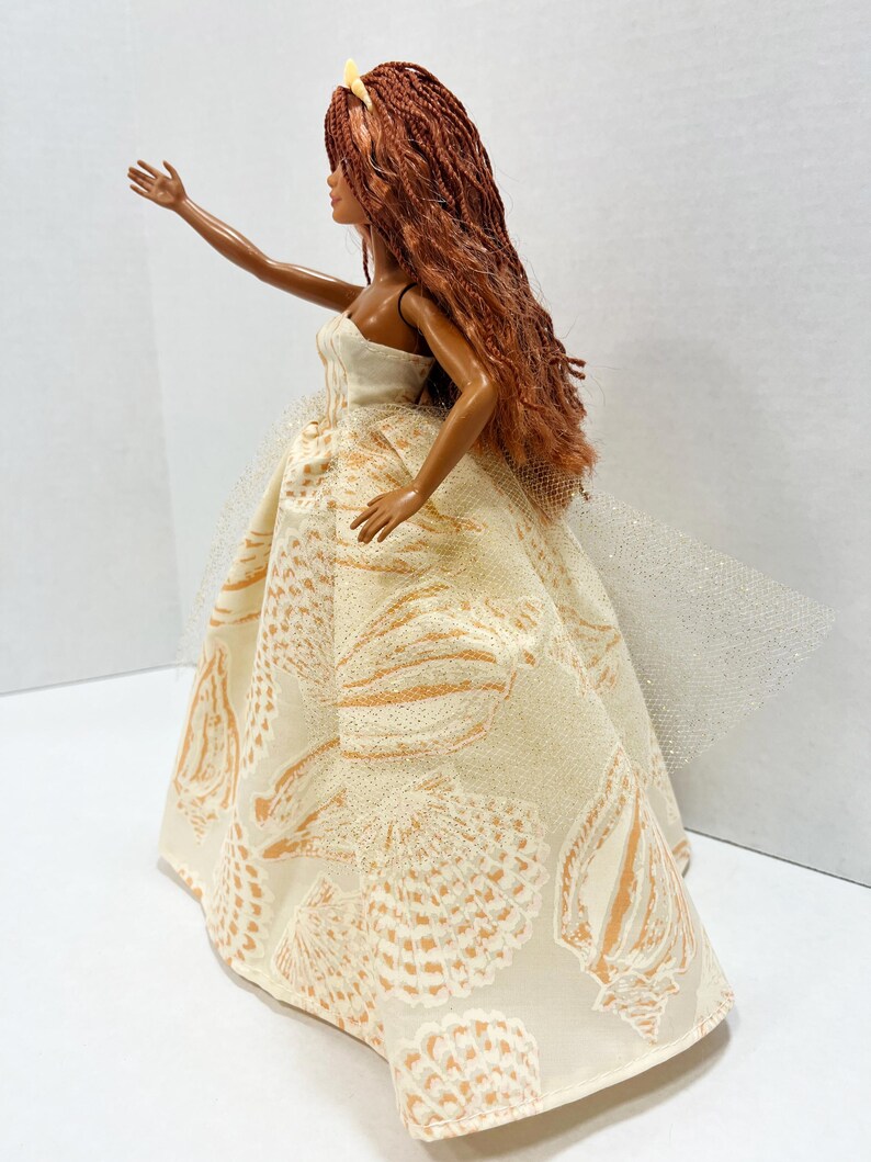 Puede incluir: Mu&ntilde;eca con largo cabello casta&ntilde;o rojizo y un vestido sin tirantes color crema. El vestido presenta un estampado de conchas en tonos dorados. Una tela transparente y brillante se extiende desde la espalda del vestido.