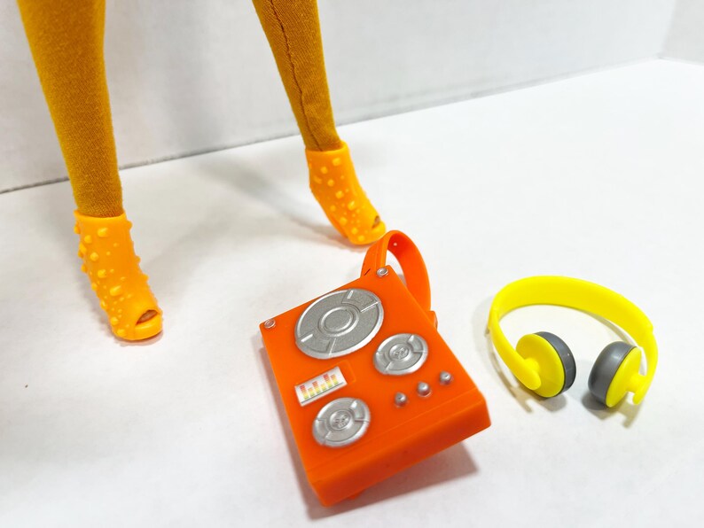 K&ouml;nnte beinhalten: Ein Paar senfgelbe Leggings, orangefarbene, genoppte Peeptoe-Schuhe, ein orangefarbenes DJ-Mischpult aus Kunststoff mit silbernen runden Drehreglern und einem kleinen rechteckigen Display sowie ein leuchtend gelber Kopfh&ouml;rer mit grauen Ohrpolstern.