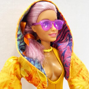 K&ouml;nnte beinhalten: Eine Puppe mit hellviolettem Haar tr&auml;gt eine durchscheinende violette Sonnenbrille, ein gelbes Halsband und eine gelbe Kapuzenjacke. Die Jacke hat ein Batikmuster auf der Au&szlig;enseite und ein buntes Futter mit tropischem Blattmuster in der Kapuze.