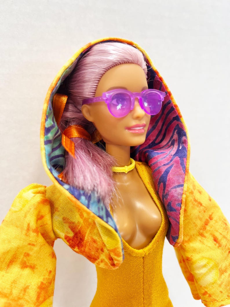 K&ouml;nnte beinhalten: Eine Puppe mit hellviolettem Haar tr&auml;gt eine durchscheinende violette Sonnenbrille, ein gelbes Halsband und eine gelbe Kapuzenjacke. Die Jacke hat ein Batikmuster auf der Au&szlig;enseite und ein buntes Futter mit tropischem Blattmuster in der Kapuze.