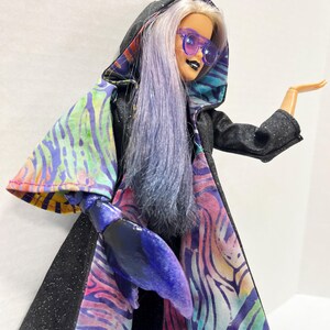 May include: A doll with long, lavender hair and purple sunglasses, wearing a black hooded cape with a colorful, patterned lining. The doll has a black lipstick and a blue lobster claw hand.