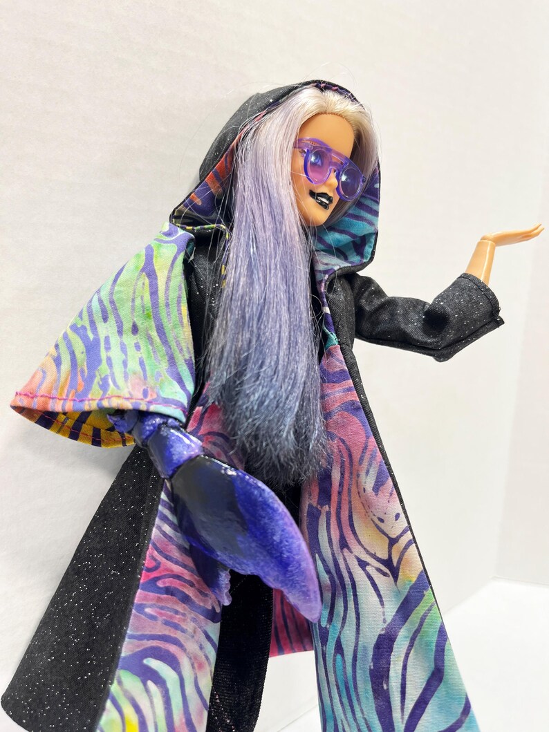 May include: A doll with long, lavender hair and purple sunglasses, wearing a black hooded cape with a colorful, patterned lining. The doll has a black lipstick and a blue lobster claw hand.