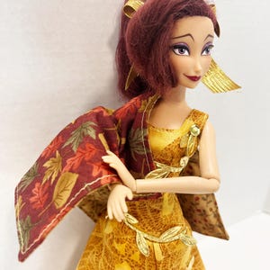 May include: A doll with dark red hair styled in a high ponytail, wearing a gold dress with a leaf pattern and a matching cape featuring a red and brown autumn leaf print. The doll has purple eyeshadow, dark red lipstick, and gold leaf-shaped accents.