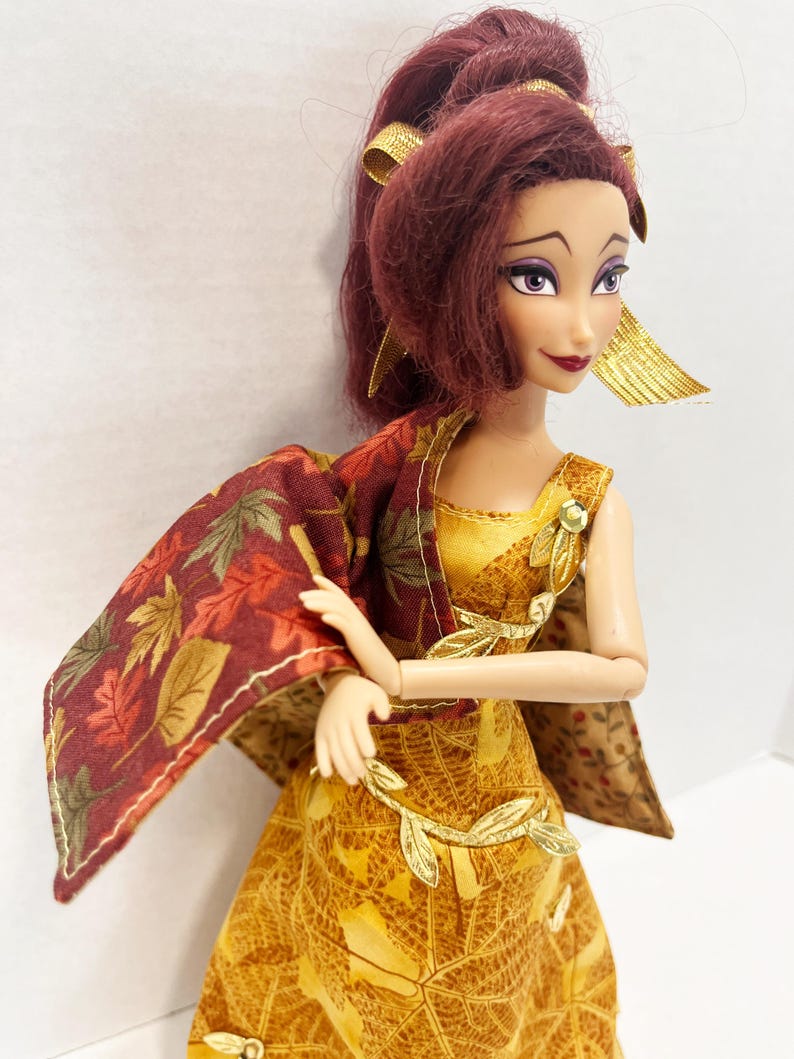 May include: A doll with dark red hair styled in a high ponytail, wearing a gold dress with a leaf pattern and a matching cape featuring a red and brown autumn leaf print. The doll has purple eyeshadow, dark red lipstick, and gold leaf-shaped accents.