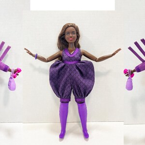 May include: A fashion doll in a purple outfit with a matching hat and accessories. The doll is wearing a purple jacket, a dress with a unique pattern, and purple boots. The doll is holding a small purse and weights. The hat has a pink flower.