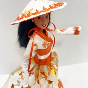 May include: A doll with long black hair wearing a white conical hat with orange leaf embroidery, a white long-sleeved top with orange leaf patterns, an orange sash, and a full skirt featuring a white and orange metallic leaf print design.