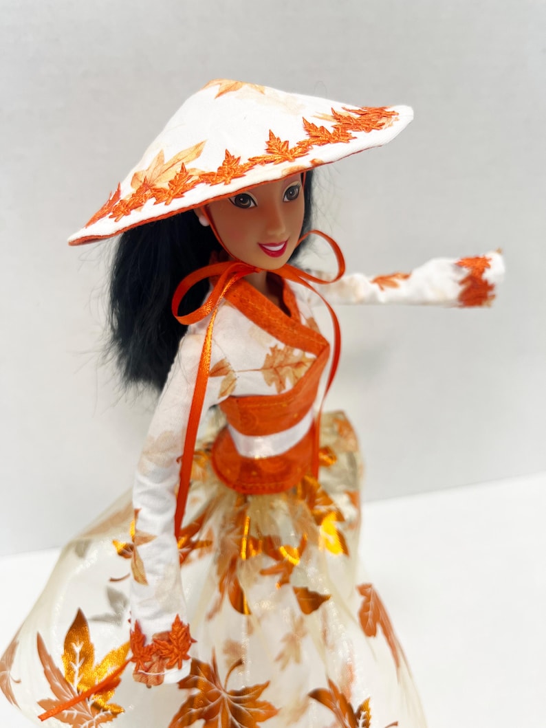 May include: A doll with long black hair wearing a white conical hat with orange leaf embroidery, a white long-sleeved top with orange leaf patterns, an orange sash, and a full skirt featuring a white and orange metallic leaf print design.