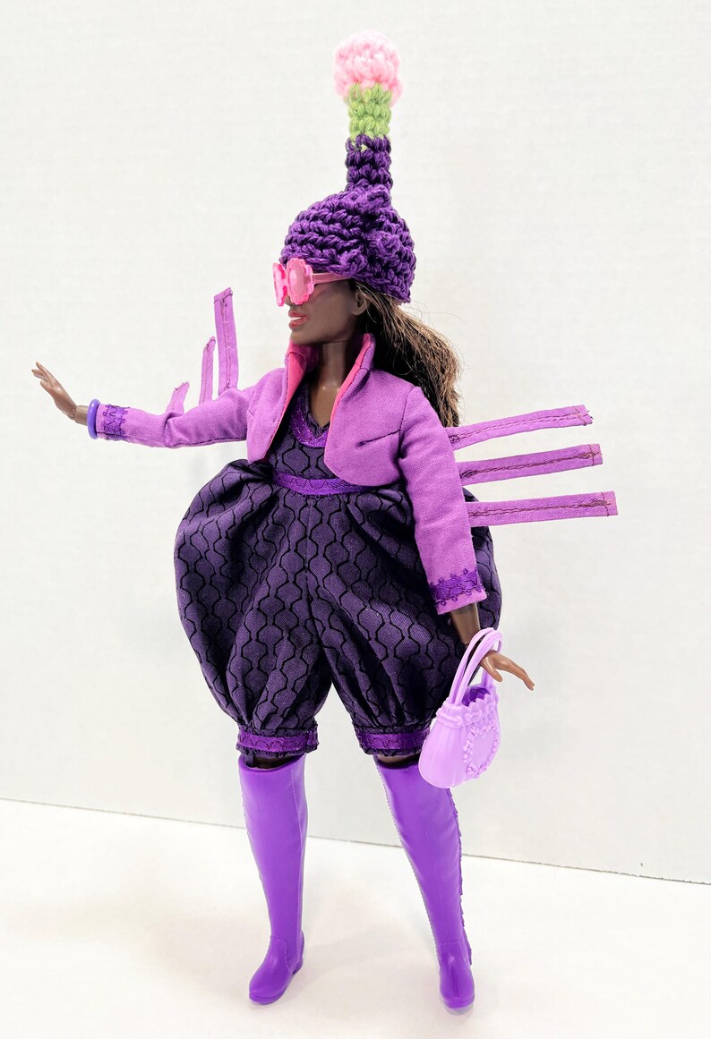May include: A fashion doll in a purple outfit, including a jacket, romper, and boots. The doll wears a unique hat with a pink and green top, pink sunglasses, and carries a matching purse. The romper has a patterned design.