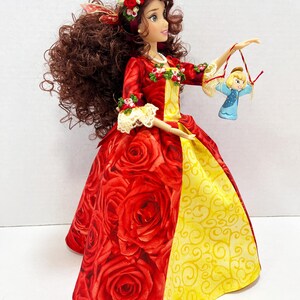 May include: A doll in a red rose-patterned gown with a yellow skirt panel and a matching floral headband. The doll holds a small ornament of a blonde girl in a blue dress. The doll has long brown curly hair and is wearing a red and yellow dress.