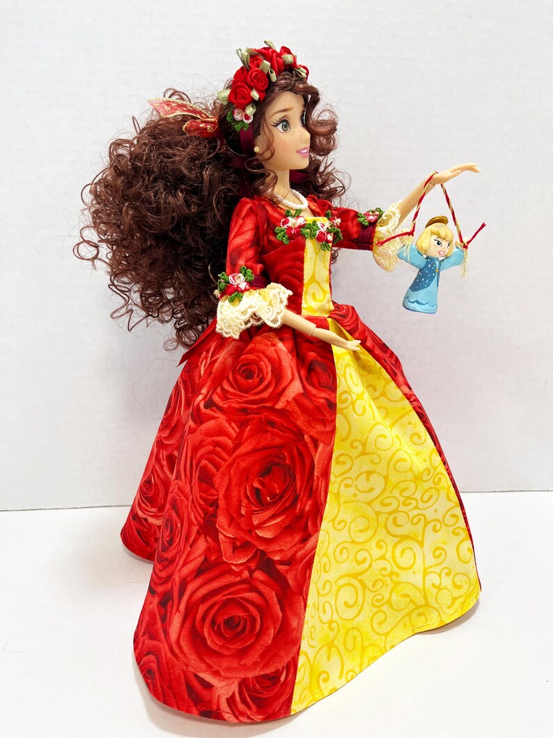 May include: A doll in a red rose-patterned gown with a yellow skirt panel and a matching floral headband. The doll holds a small ornament of a blonde girl in a blue dress. The doll has long brown curly hair and is wearing a red and yellow dress.