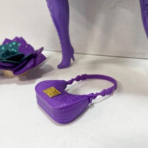 May include: A purple doll accessory set featuring a small handbag with a gold square clasp, a flower-shaped headpiece with teal and purple accents, and purple high-heeled boots. The boots have a lace-up design.