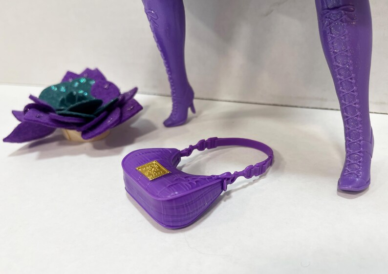 May include: A purple doll accessory set featuring a small handbag with a gold square clasp, a flower-shaped headpiece with teal and purple accents, and purple high-heeled boots. The boots have a lace-up design.