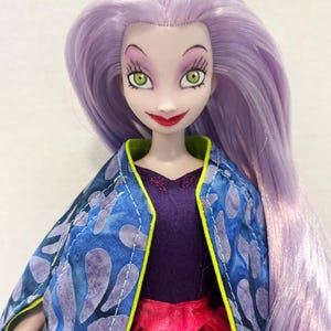 Pu&ograve; includere: Una bambola con lunghi capelli lisci color lavanda, pelle chiara e occhi verdi. La bambola indossa un top viola scuro con una gonna rossa e un mantello blu con un motivo a foglie viola e un bordo verde lime brillante sul davanti.