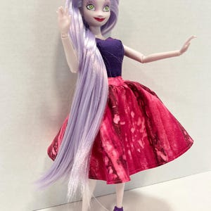 Pu&ograve; includere: Una bambola con lunghi capelli lisci color viola chiaro, occhi verdi e labbra rosse. La bambola indossa un top senza maniche viola scuro e una gonna ampia al ginocchio con un motivo marmorizzato rosso e rosa scuro, abbinata a sandali con tacco.