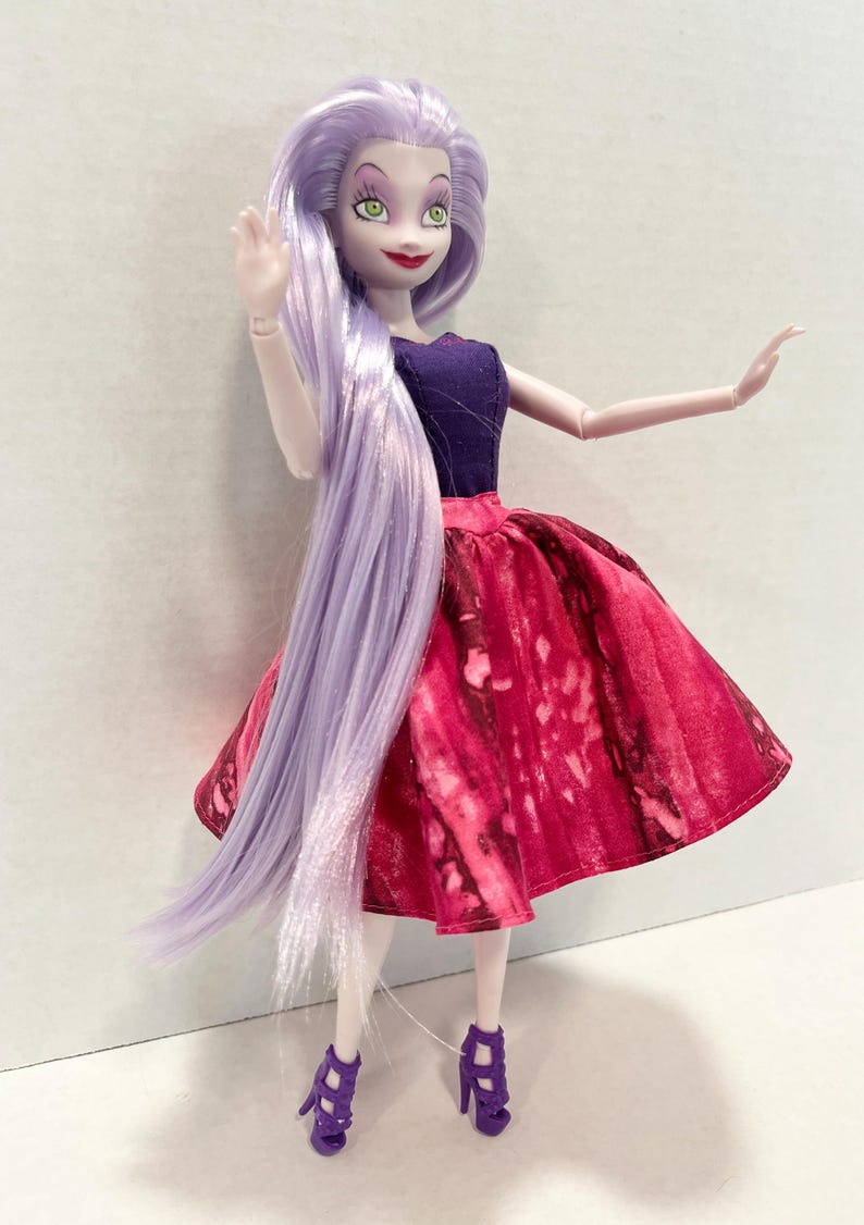 Pu&ograve; includere: Una bambola con lunghi capelli lisci color viola chiaro, occhi verdi e labbra rosse. La bambola indossa un top senza maniche viola scuro e una gonna ampia al ginocchio con un motivo marmorizzato rosso e rosa scuro, abbinata a sandali con tacco.
