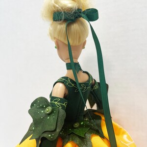 May include: A doll with blonde hair styled in an updo with a dark green ribbon. The doll wears a dark green bodice featuring embroidered leaf patterns, a matching choker, and long sleeves with layered leaf-shaped cuffs over a yellow skirt.