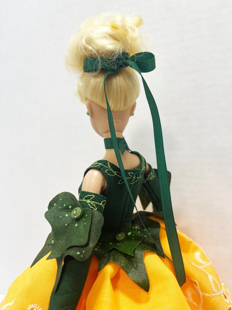 May include: A doll with blonde hair styled in an updo with a dark green ribbon. The doll wears a dark green bodice featuring embroidered leaf patterns, a matching choker, and long sleeves with layered leaf-shaped cuffs over a yellow skirt.