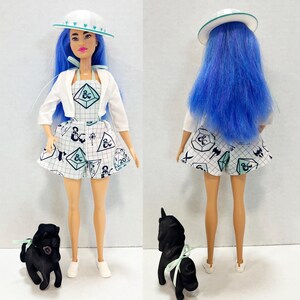 May include: A doll with long blue hair wearing a white hat with teal hearts, a white cropped jacket, and a white romper featuring a grid pattern with Dungeons and Dragons symbols. A small black dog figurine with a teal ribbon is positioned nearby.