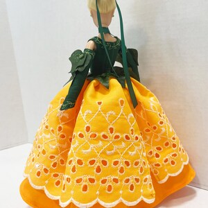 May include: A doll with blonde hair styled in an updo with a green ribbon. The doll wears a dark green bodice with leaf-like sleeves and a vibrant yellow skirt featuring white eyelet embroidery and a scalloped hemline with white stitching.