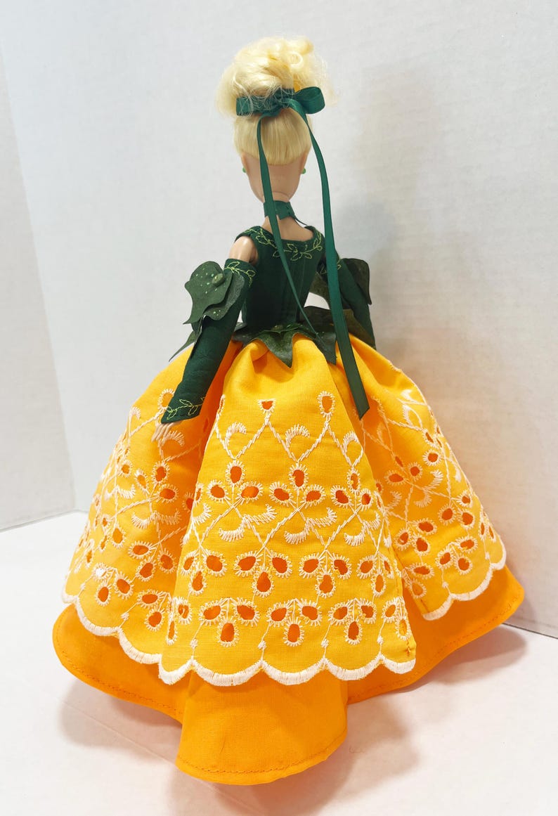 May include: A doll with blonde hair styled in an updo with a green ribbon. The doll wears a dark green bodice with leaf-like sleeves and a vibrant yellow skirt featuring white eyelet embroidery and a scalloped hemline with white stitching.