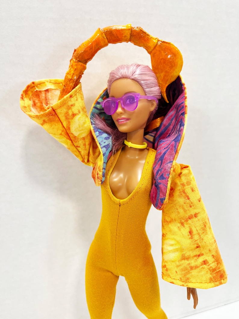 K&ouml;nnte beinhalten: Eine Puppe mit hellrosa Haar tr&auml;gt eine violette Sonnenbrille, ein gelbes Halsband und einen senfgelben Body. Die Puppe tr&auml;gt eine Kapuzenjacke mit gelb-orangefarbenem Batikmuster au&szlig;en und einem blau-violett gemusterten Innenfutter.