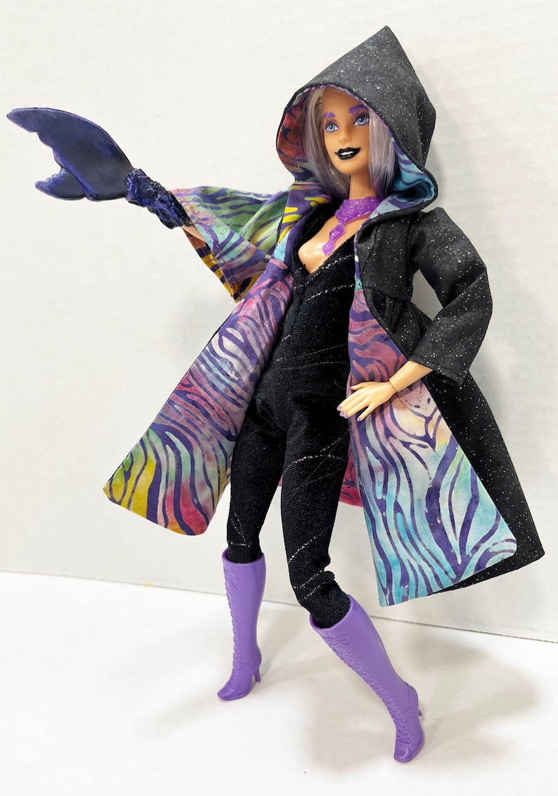 May include: A doll in a black jumpsuit and a hooded black coat with a colorful patterned lining. The doll has purple boots and a purple necklace. A large, dark blue, claw-like accessory extends from the doll's arm.