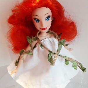 Puede incluir: Una mu&ntilde;eca de pelo rojo con un vestido blanco con hojas verdes y cuerda alrededor de la cintura. La mu&ntilde;eca tiene ojos azules y una cara sonriente.